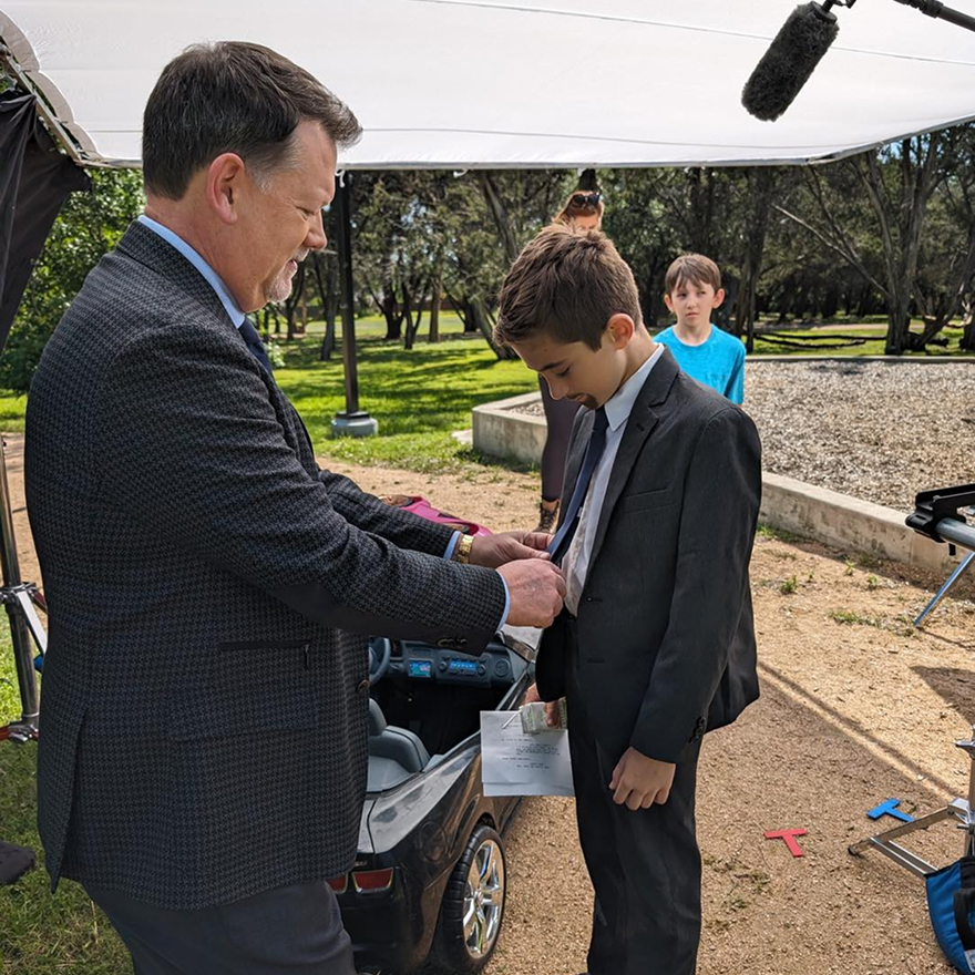 Attorney Dean Boyd helping a young boy adjust his tie during an outdoor community event.