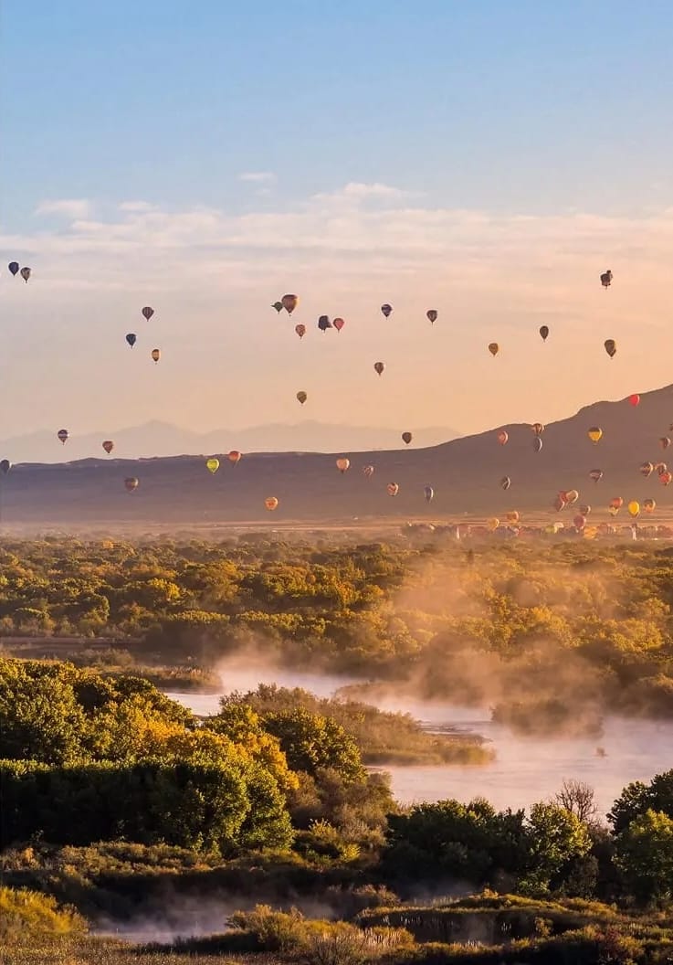 Hot air balloons rise over a misty river, autumn trees, and mountains in Albuquerque, NM