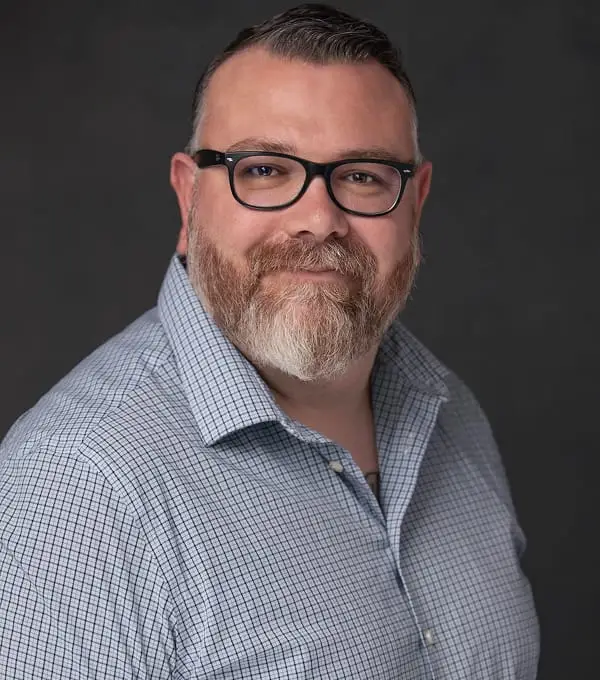 Smiling man with beard, glasses, in checkered shirt for a headshot.